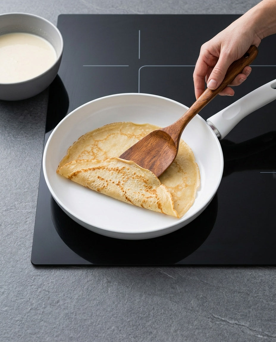 La spatule plate en acacia glisse sous une crêpe dans une poêle en céramique blanche
